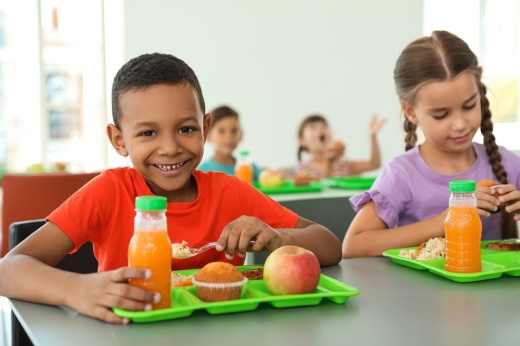 Children sit at a lunch table with meals on green trays.