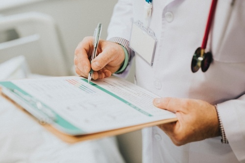 This is a close up of a person holding a clipboard and a pen who is wearing a white coat and a stethoscope. 
