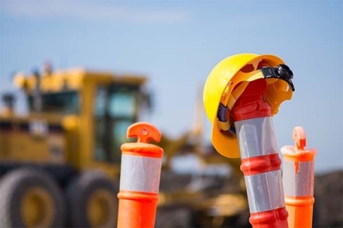 Close up of the top of traffic cones, with a construction hat on one of the cones. A construction vehicle is in the background.