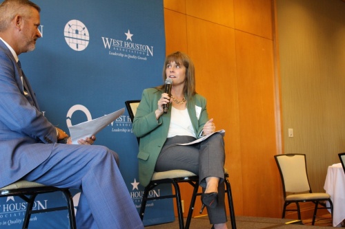 A woman in a green blazer and gray pants holds a microphone and is sitting next to a man in a blue suit.
