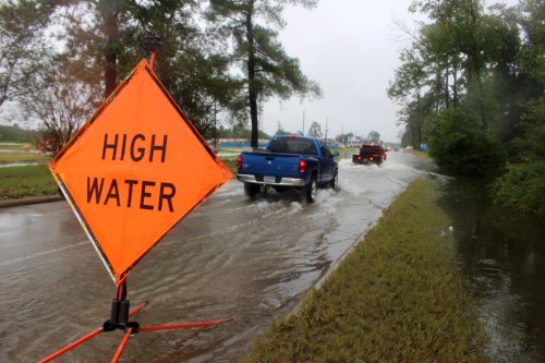 An orange sign indicating high water is atop a road with standing water, and a blue truck is driving through the water.