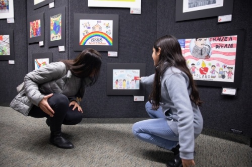 Nora Rotem, elementary school art finalist in the Martin Luther King Jr. diversity competition, shows her mother, Ori Rotem, her piece. Finalist and semifinalist art pieces filled the common area of Williams High School. (Liesbeth Powers/Community Impact Newspaper)