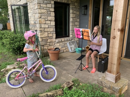 A photo of a girl playing a saxaphone on a porch with another girl holding a bike and watching