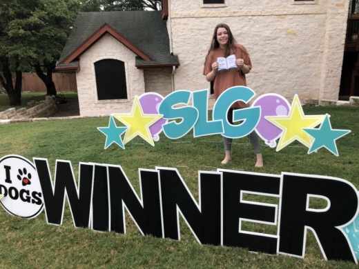 A photo of a student, Kaylin Whitaker, holding up a check behind a yard sign that reads "SLG Winner."