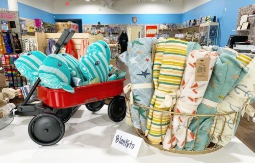 A basket of baby blankets and a red wagon with five small stuffed whales in it, both on a display shelf