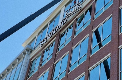 A crane lowers a a large bank sign into place on the side of a building. 