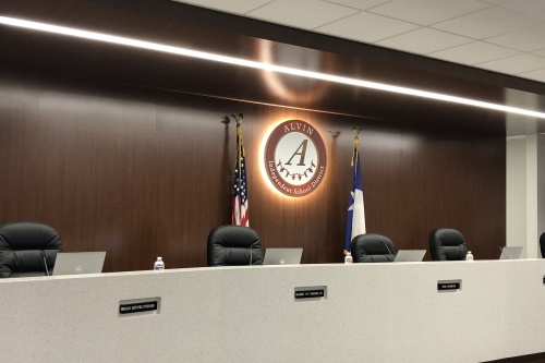 The Alvin ISD board room, four chairs behind a desk with four laptops in front of the American and Texas flags.
