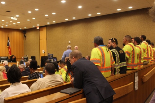 People in yellow safety vests stand at the back of an orange courtroom. Another man in a black suit leans on the courtroom benches.