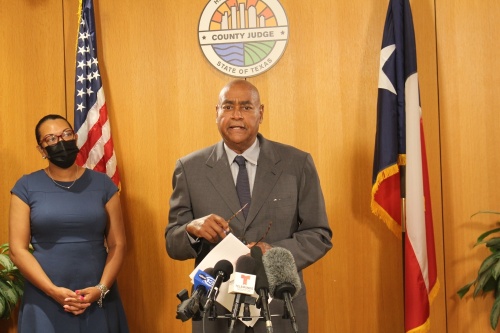 A man with glasses in a gray suit and tie gives a speech in front of a podium. A woman wearing a face mask stands behind him in a blue dress. They stand in front of a wooden orange wall and the American and Texas flags.