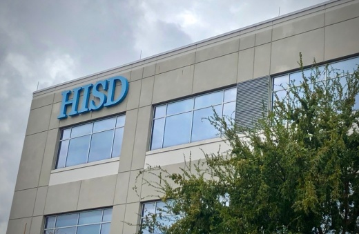 A close-up of a gray concrete building with a blue HISD sign in the corner, along with a tree in the bottom right of the grame.
