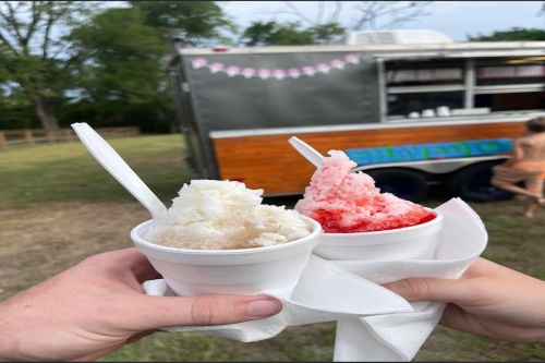 Two white hands holding a white shaved-ice and a red shaved-ice each. A brown, black and blue trailer is visible in the background