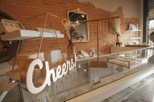 In silver text, a sign saying "Cheers" on top of a glass and metal case against a brown wall featuring various kinds of jewelry