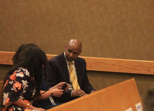 A man in a black suit with a gold tie talks to two women on the benches of an orange county courtroom.