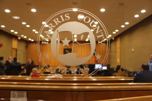 A white logo on a glass window saying "Harris County Texas" on a door to the courtroom is in the foreground, and there is an orange courtroom visible in the background behind the glass with at least a dozen people sitting or standing in the room.