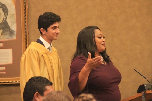 A high school student wears a gold graduation robe next to a woman in a burgundy dress. 