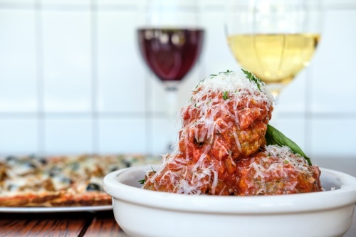 bowl with meatballs and two glasses of wine and a pizza behind it