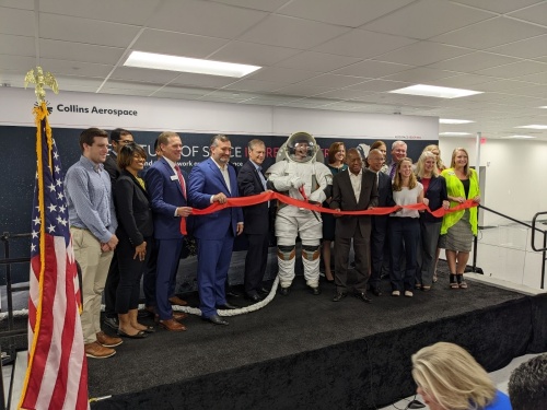 People and a man in a space suit hold up a ribbon for a ribbon cutting ceremony