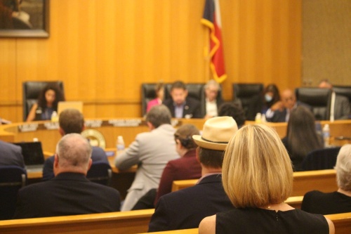 A woman with short blond hair sits at the back of an orange courtroom behind other court attendees as the members of the court have discussions.
