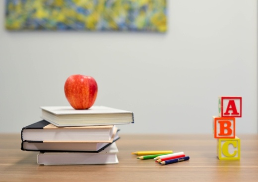 A red apple sits on a stack of books next to some colorful letter blocks and colored pencils on a table.