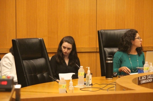 An empty leather chair is next to a woman with glasses in a green blouse. Another woman in a black shirt is sitting behind the chair.