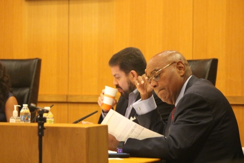 A man wearing glasses looks down at a piece of paper while a man with a beard next to him takes a sip of his drink.