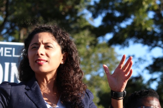 A woman with curly brown hair wearing a navy suit jacket speaks outside at a voter rally.
