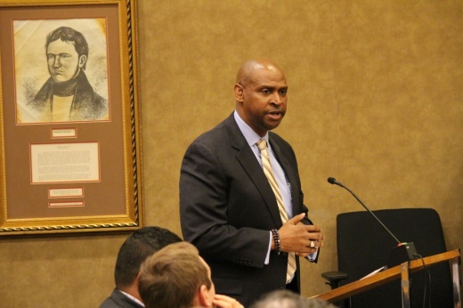 A man in a black suit speaks in front of a podium in a courtroom.
