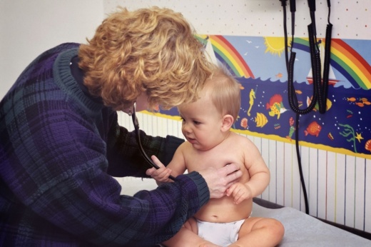 A curly-haired adult checks the heartbeat of a blond child wearing a diaper.