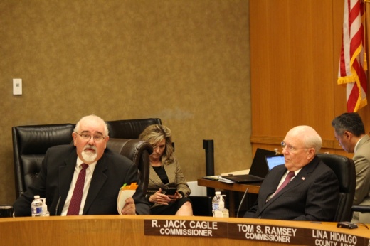 Two men wearing glasses and suits sit next to each other. The man on the left is holding a file as he speaks. A woman and a man are behind the two men doing work on a computer.