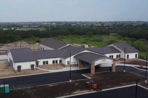 A church building under construction sits in front of a green landscape and blue sky in Cedar Park. The building has a gray roof and has four different wings facing out in different directions North, East, South and West.