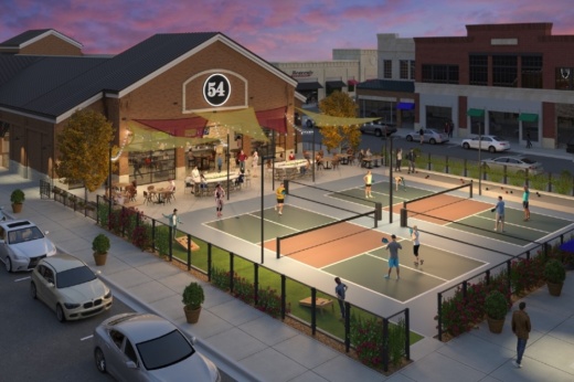 Two pickleball courts sit in front of a restaurant with outdoor seating. The front of the restaurant has a circular black sign that says 54 with white font. A street lined with parked cars sits adjacent to the restaurant and pickleball courts.