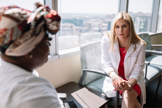 A female doctor seated on the right speaks with a woman on the left seen in the foreground from the side.