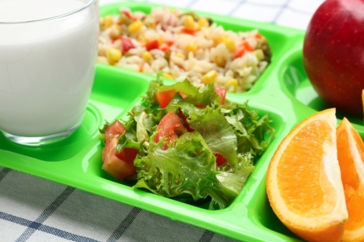 Stock image shows a lunch tray with food, including an apple, orange slices, salad and a glass of milk.