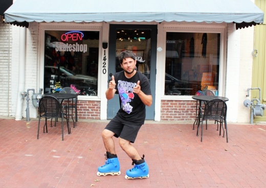 a man stands outside a storefront wearing roller blades giving two thumbs up with his pointer fingers out