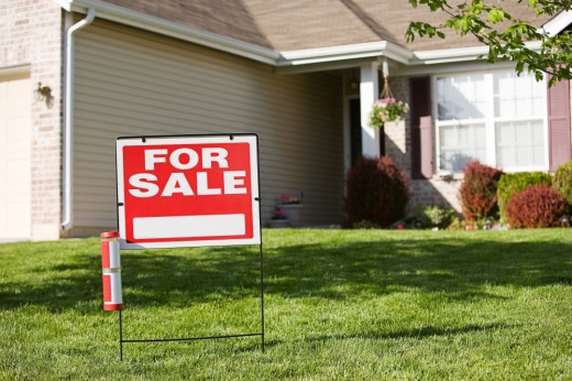 In front of a tan house, a red sign says 'for sale' in white lettering.
