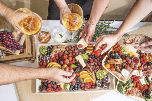 Hands reach across colorful charcuterie boards featuring fruit and meats.