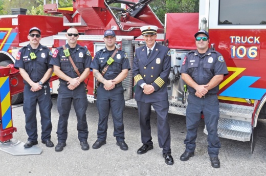 A line of firefighters stands in front of a fire engine.