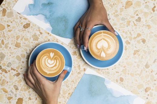 An overhead view of two hands holding two blue coffee cups with swirled foam on top.