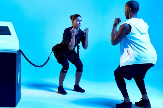 A trainer leads a woman wearing an electro-muscle stimulation vest in a squat.