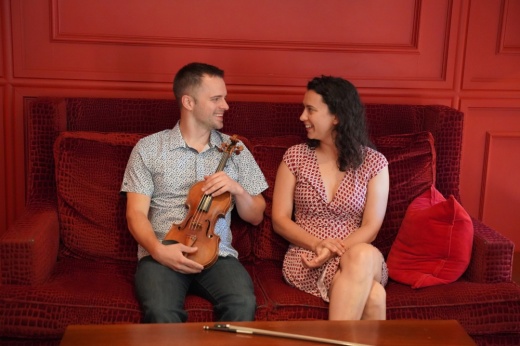ATX Music Co-Founder Geoffrey Herd and Co-Artistic Director Anna Petrova sit next to each other on a red sofa. Herd holds his violin in his lap.