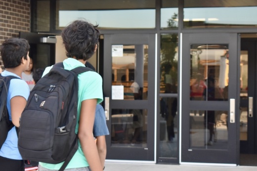two kids with backpacks on walk toward a school's door
