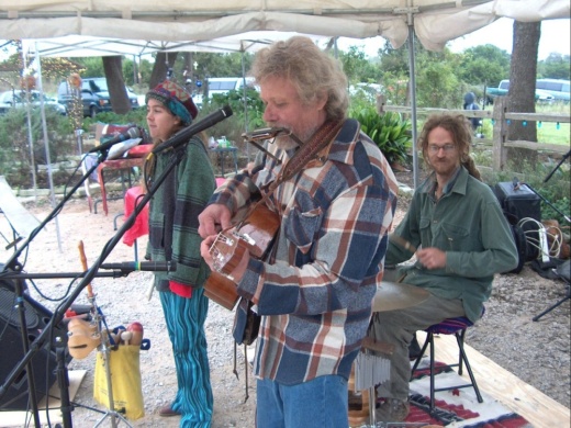 hans flentge plays a guitar and a harmonica with two other musicians on a stage