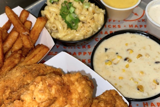 A plate of fried chicken is shown on a table with white cheddar mac & cheese and roasted creamed corn, fries, and two dipping sauces.