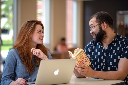 The photo shows two students studying with a book and a laptop.