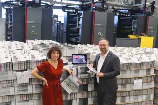 community impact vice president of sales and marketing stands next to ceo john garrett in the printing press in front of piles of newspapers and a laptop with the CI 360 logo on the screen