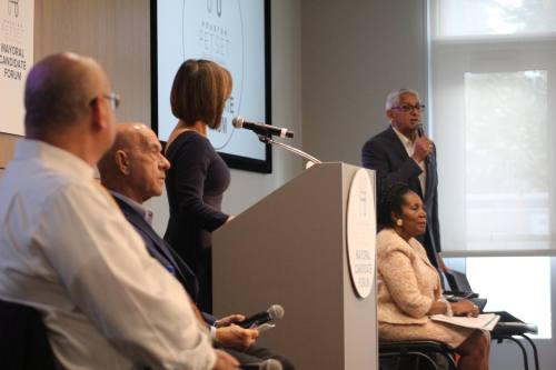 three houston mayor candidates sit in chairs while one candidate stands at a forum with a moderator at a podium in the center