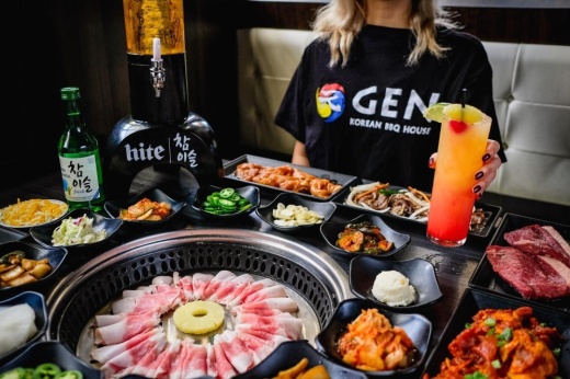 a person holds a cocktail while seated at a table covered in Korean barbecue dishes