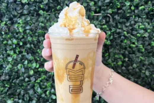 A caramel frappe is held up by a person's hand in front of a wall of greenery outside.