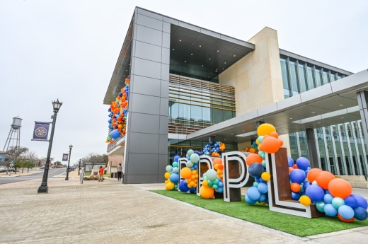 The front of a public library is shown on its grand opening with balloons decorating the side of the building and signage.