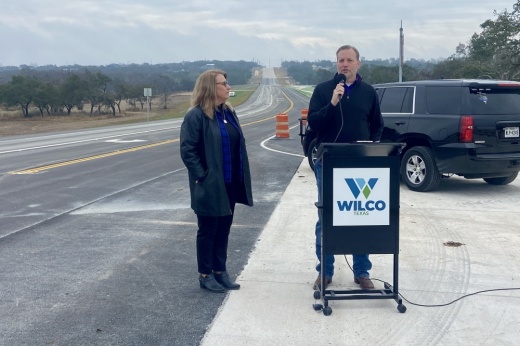 A woman and man speak at a podium in front of a road.